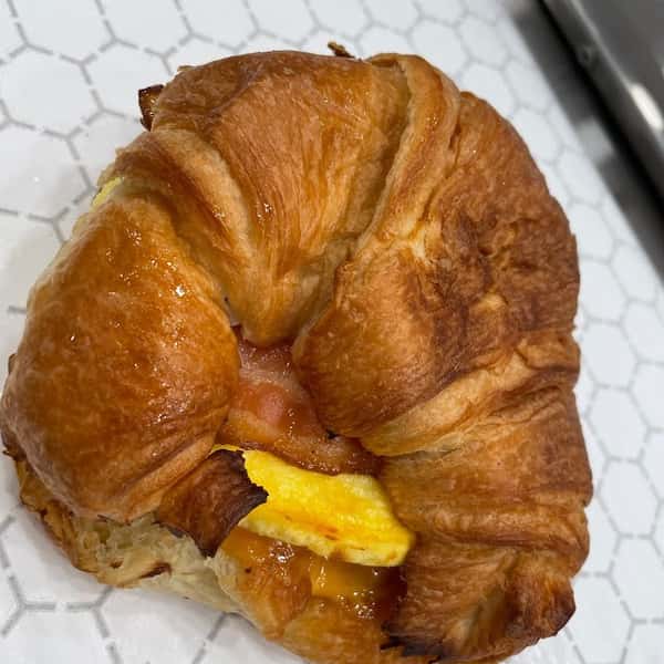 Toasted croissant breakfast sandwich filled with fluffy scrambled eggs, crispy bacon, and melted cheese, served on a white patterned liner.
