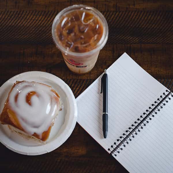 Top-down view of a classic cinnamon roll with icing and an iced coffee from Liberty Brew, next to a spiral notebook and pen on a wooden table.