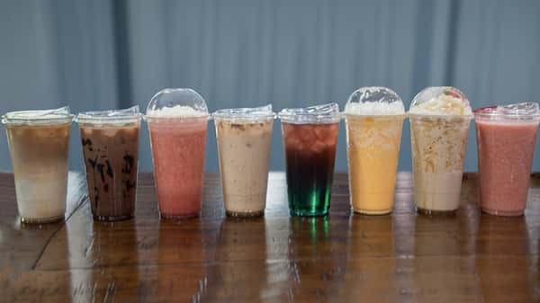 Lineup of eight colorful Liberty Brew specialty drinks in clear cups, showcasing a vibrant variety of iced coffees, fruit smoothies, and blended beverages on a rustic wooden table.