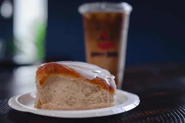 Freshly baked cinnamon roll with thick vanilla icing on a paper plate, paired with a chilled iced coffee from Liberty Brew, served on a rustic wooden table.
