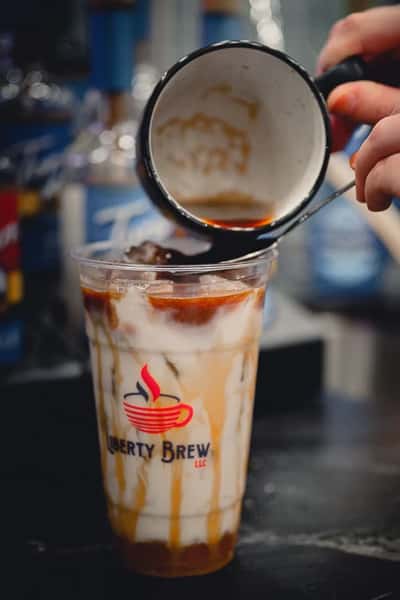 Barista pouring rich espresso over an iced caramel macchiato at Liberty Brew, creating a creamy swirl in a clear plastic cup with branded logo.