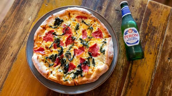 Pizza with white cheese, red salami, and dark greens on a plate next to a green beer bottle on a wooden table.