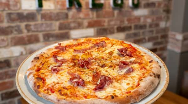 Salami pizza with melted cheese and red sauce on a white plate, with a brick wall and "PREGO" sign in the background.
