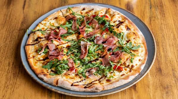 Pizza with white sauce, mozzarella, arugula, prosciutto, and balsamic glaze on a grey plate.