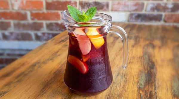 Red sangria in a clear pitcher with mint, lemon, apple slices, and ice on a wooden table.