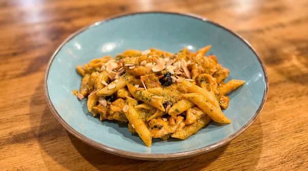 Penne pasta with creamy orange-green sauce, chicken, and sliced almonds in a teal bowl on a wooden table.