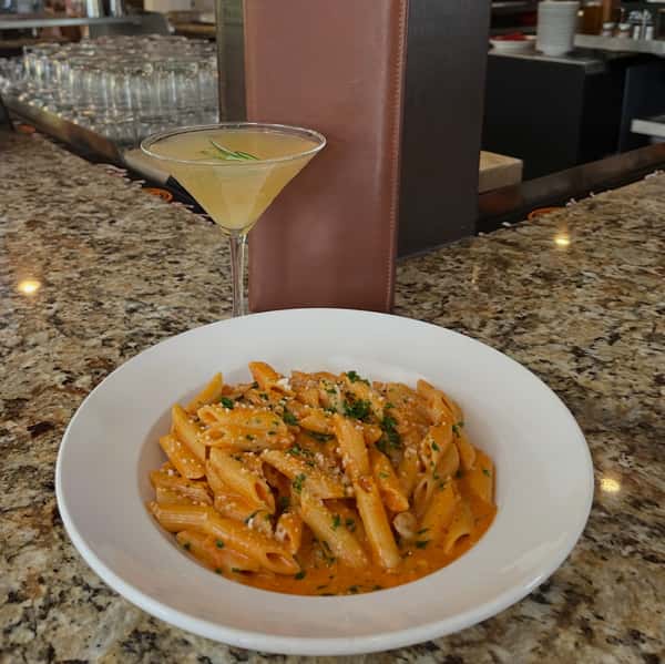 Penne pasta dish on a plate with a cocktail on a tabletop