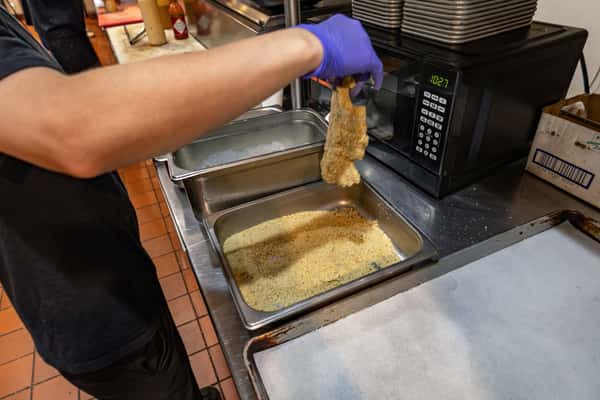 Chef preparing breaded chicken in a commercial kitchen, dipping fillets into seasoned breadcrumbs for frying or baking