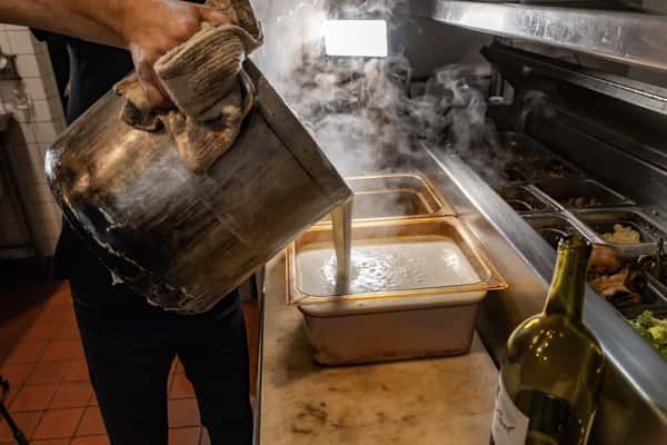 Chef pouring freshly made Alfredo sauce into warming pans in a busy kitchen, with steam rising from the rich, creamy sauce.