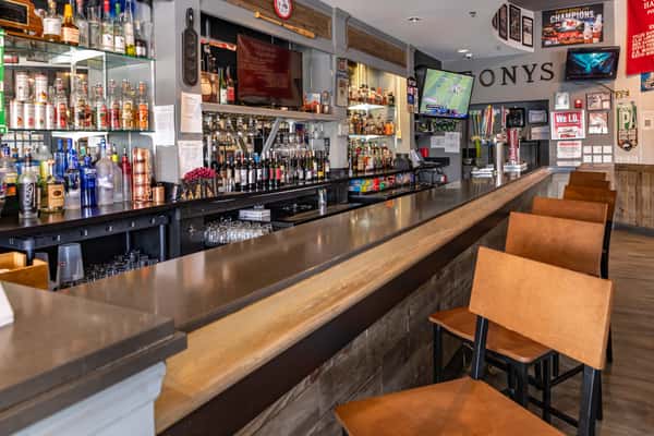Modern sports bar with a sleek wooden countertop, high-top seating, and a fully stocked liquor display, featuring multiple flat-screen TVs showing live games and sports memorabilia on the walls.