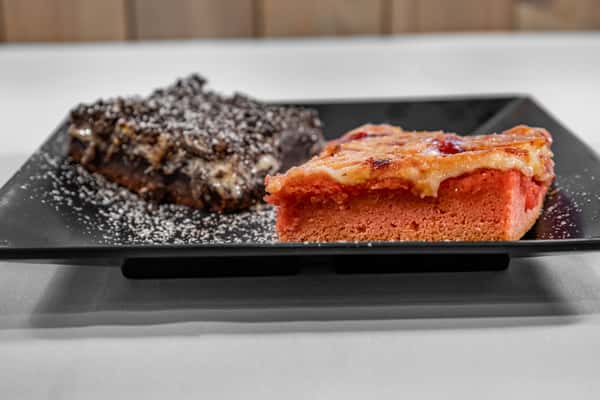 Plate of assorted desserts featuring a slice of gooey chocolate brownie and a square of fruity cobbler, dusted with powdered sugar and served on a black square plate.