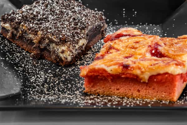 Close-up of decadent desserts including a rich chocolate brownie with powdered sugar and a slice of fruit-topped pink cobbler on a black plate