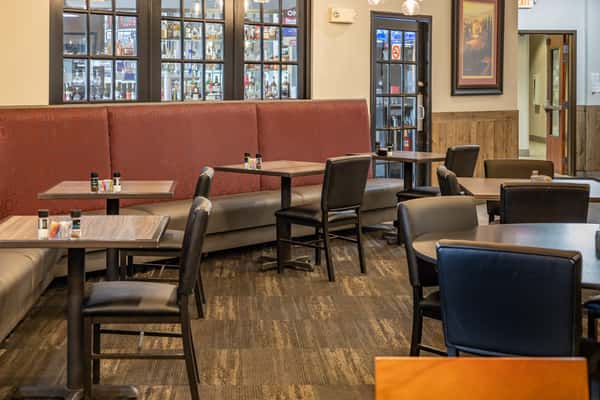 Cozy dining area with red upholstered booth seating, wooden tables, and black chairs, set against large windows and warm, neutral decor.