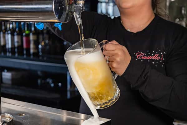 Server pouring a frothy mug of draft beer behind the bar at Castello's, capturing a fresh, cold pour overflowing with foam.