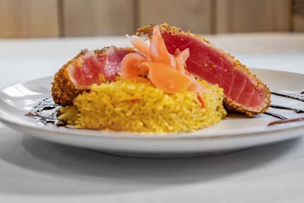 Side view of sesame-crusted ahi tuna, sliced and served rare with yellow seasoned rice and pickled ginger, accented by a balsamic glaze swirl.