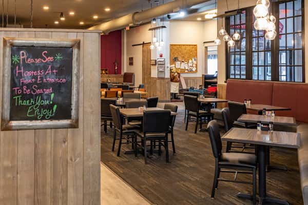Inviting dining area with wooden tables, cushioned booths, and a chalkboard sign directing guests to the hostess stand, enhanced by warm lighting and rustic decor.