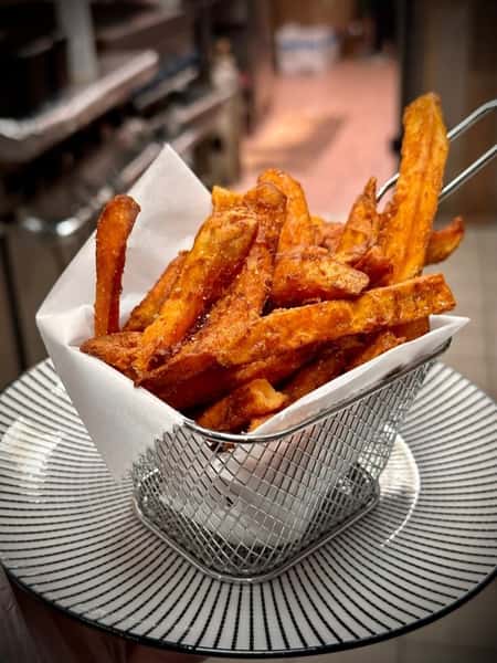 Crispy sweet potato fries served in a metal basket lined with parchment paper, presented on a stylish black and white plate.