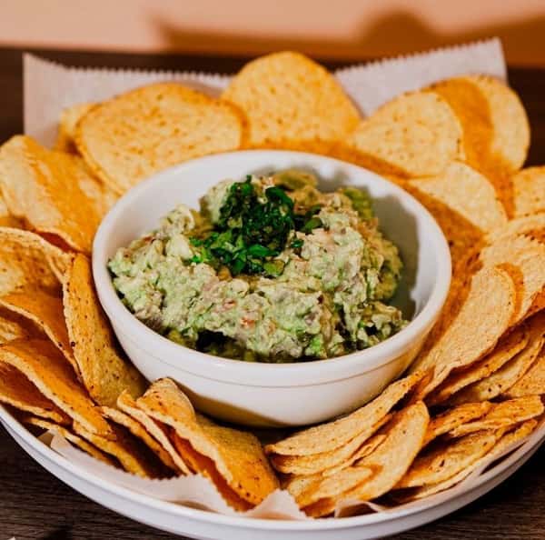 Creamy guacamole topped with chopped cilantro, served in a white bowl surrounded by golden tortilla chips on a white plate.