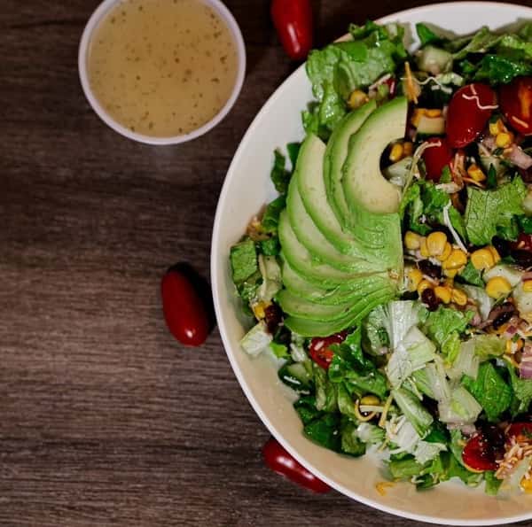 Southwest avocado salad topped with sliced avocado, roasted corn, black beans, shredded cheese, cherry tomatoes, and romaine lettuce, served with a side of vinaigrette dressing.