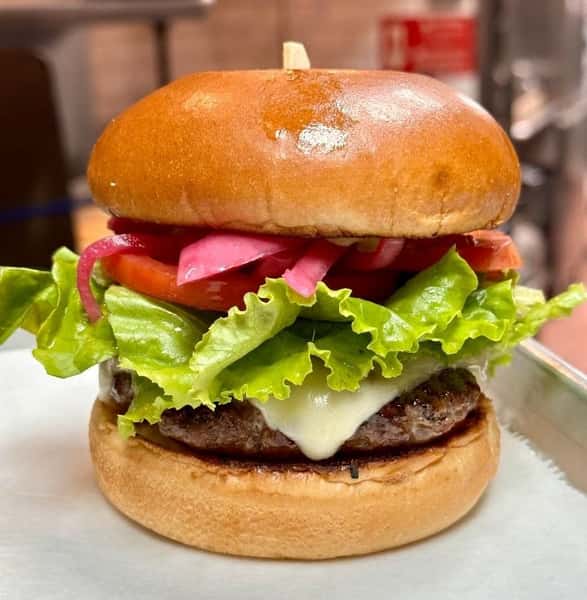 Gourmet cheeseburger topped with white cheddar, leaf lettuce, tomato, and pickled red onions, all in a glossy toasted bun.
