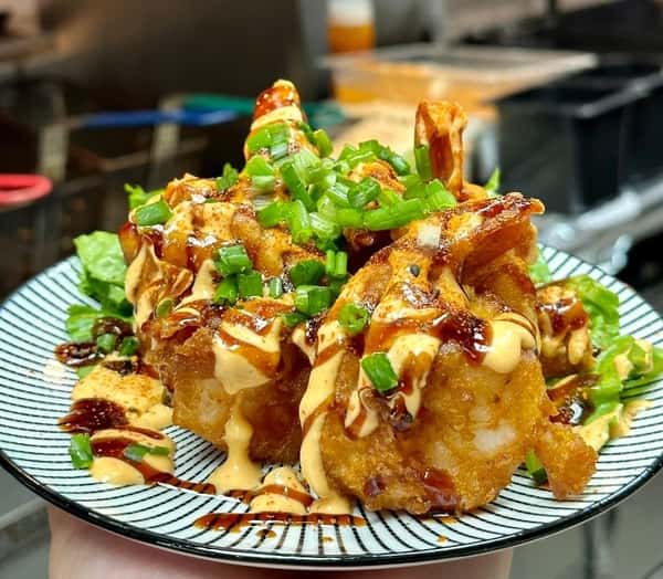 Crispy fried shrimp drizzled with spicy mayo and sweet soy glaze, garnished with chopped green onions and served over a bed of lettuce on a black-striped plate.