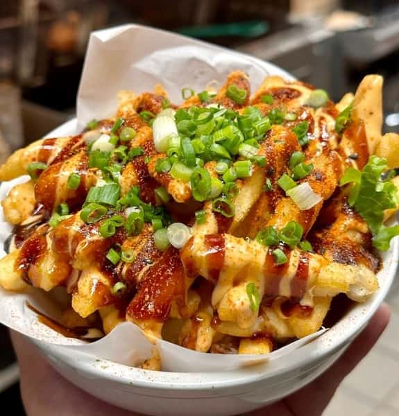 Loaded fries drizzled with spicy mayo and BBQ sauce, topped with chopped green onions and served in a white bowl lined with parchment paper.