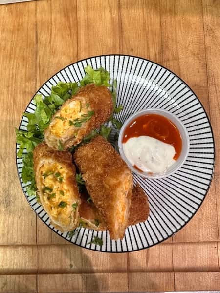 Halved buffalo chicken egg rolls with a crispy golden crust, filled with shredded chicken and cheese, served with a duo of ranch and hot sauce on the side.