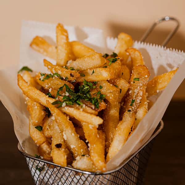 A generous serving of crispy, golden-brown french fries seasoned with fresh herbs and served in a classic metal basket at The Local Press in Ozone Park, representing a signature comfort food side dish.