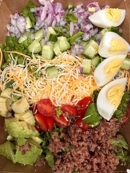 Overhead view of a loaded taco salad with seasoned ground beef, hard-boiled eggs, shredded cheese, cherry tomatoes, avocado slices, diced cucumber, red onion, and chopped romaine lettuce, arranged in neat rows.