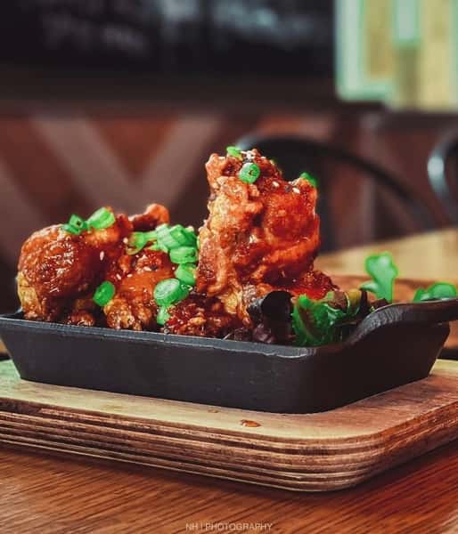 Korean-style fried chicken wings glazed in spicy sauce and topped with fresh scallions, served in a cast iron dish over a wooden board.