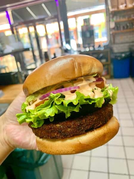 Handheld plant-based burger layered with a crispy veggie patty, leaf lettuce, red onions, pickles, and creamy sauce on a soft brioche bun.