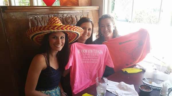 three females posing for picture. one is wearing a hat, other two are holding up shirts