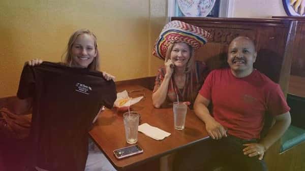 three people smiling sitting a a table. one person is wearing a hat