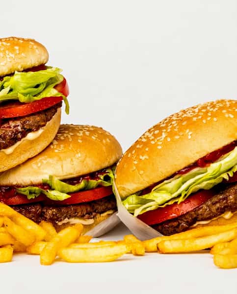 Three burgers with beef patties, lettuce, tomato, and sesame seed buns, partially wrapped in white paper, next to french fries.