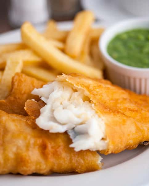 Fish and chips with golden-brown battered fish, showing flaky white fish inside, alongside thick-cut fries and a small dish of green pea mash.