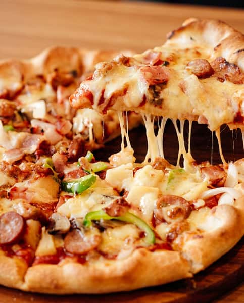 Pizza slice lifted from a whole pizza, with melted cheese stretching, topped with sausage, bacon, green peppers, and onions.