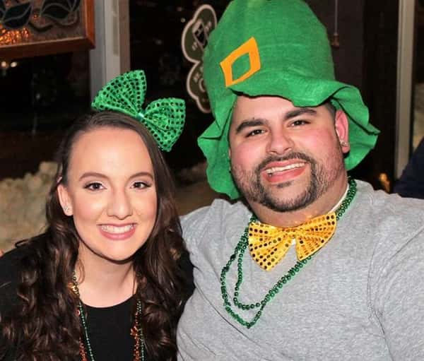 Male and female smiling in photo. female is wearing a bow, male is wearing large hat, bow tie, and beads around his neck
