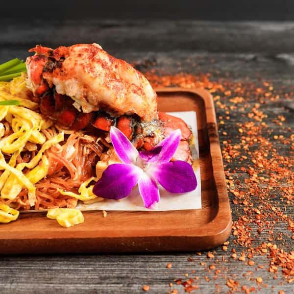 gourmet lobster pad thai with stir-fried noodles, egg, and fresh herbs garnished with an orchid on a wooden serving tray