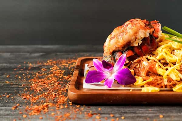 pad thai with lobster tail on wooden tray garnished with edible orchid and chili flakes, authentic Thai noodle dish presentation