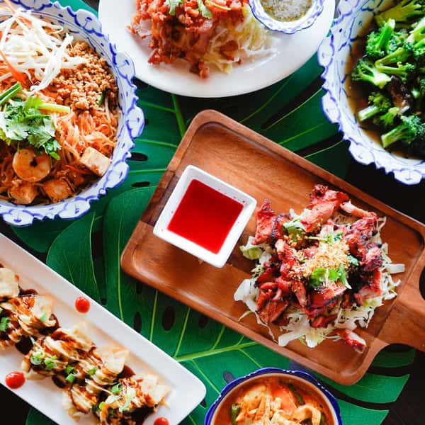 assorted Thai dishes including pad thai, grilled chicken, stir-fried broccoli, and dumplings with dipping sauces on a vibrant tropical table setting