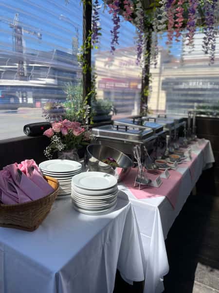 restaurant buffet setup with chafing dishes, stacked plates, and pink napkins on a decorated table with floral accents and natural light