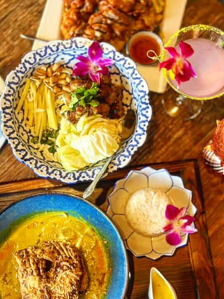 colorful Thai food spread featuring khao soi curry, fresh mango salad, jasmine rice, and a pink cocktail garnished with orchids on a wooden table