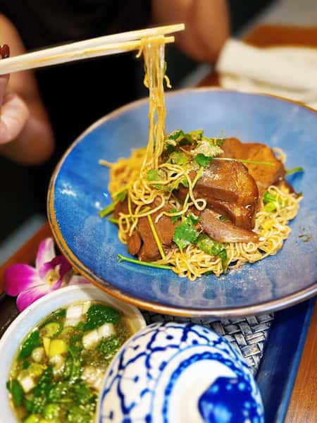 Thai egg noodle bowl with braised pork belly, fresh herbs, and chili dipping sauce served in a blue ceramic dish