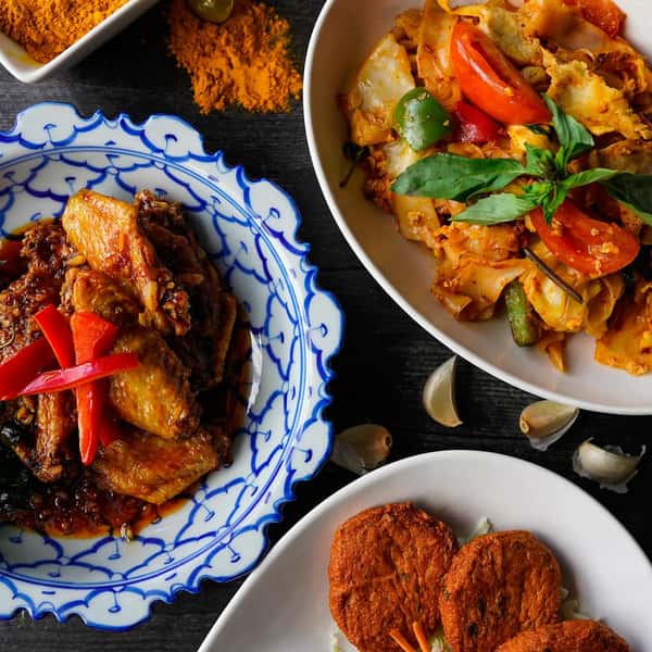 Thai drunken noodles (pad kee mao) with shrimp, bell peppers, and fresh basil alongside spicy glazed chicken wings and crispy Thai fish cakes on a dark wood table