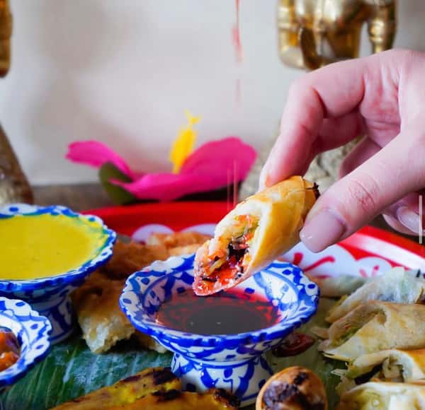 crispy spring rolls being dipped in sweet chili sauce with assorted Thai appetizers and dipping sauces on a vibrant platter