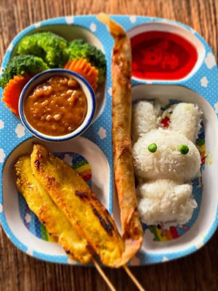 kid-friendly Thai meal with chicken satay, spring roll, peanut sauce, fresh vegetables, and rice shaped like a bunny with dipping sauces