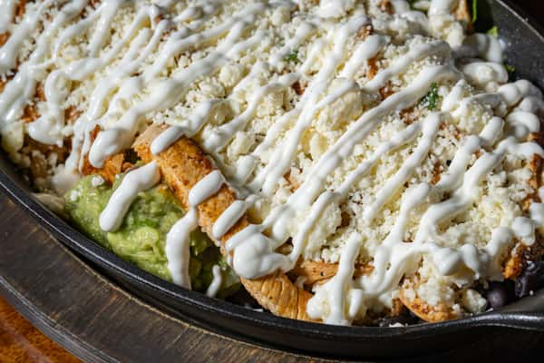 Grilled chicken enchiladas topped with crumbled cheese, crema drizzle, and guacamole on a hot skillet.