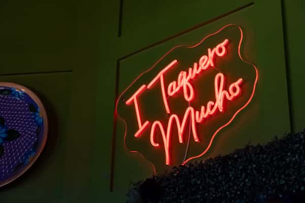 Red neon sign reading "I Taquero Mucho" on a dark green wall beside a decorative blue plate.
