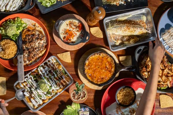 Assorted Mexican dishes including enchiladas, queso dip, and guacamole fries served on a wooden table with tortilla chips.