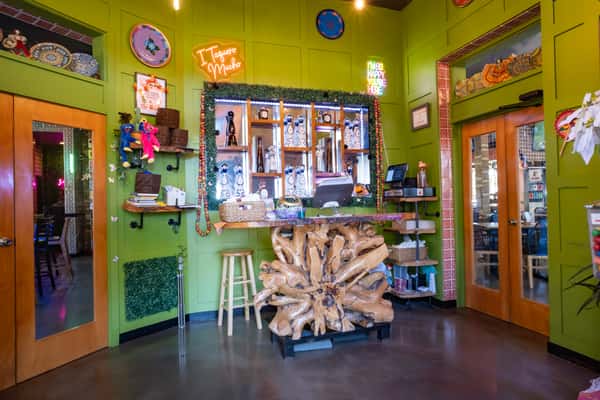 Vibrant restaurant check-in counter with neon "I Taquero Mucho" and "I'm So Happy You're Here" signs, tequila bottles, and playful decor.